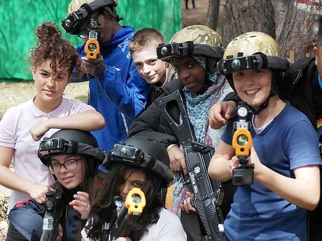 Groupe d'ados et enfants portant des casques et un fusil faisant une partie de laser game extérieur en colonie de vacances d'été