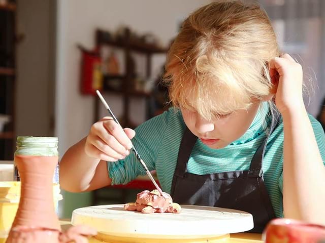 Vue sur une jeune qui fait de la poterie et qui peint sa réalisation lors d'une colo de vacances dédiée aux Beaux Arts