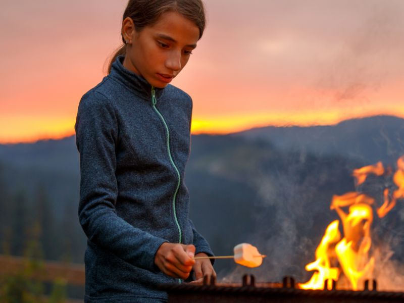 Jeune enfant qui fait fondre son chamallow sur le feu de camp en colo de vacances cet été