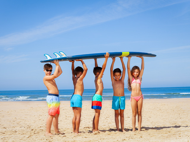 jeunes enfants tenant une planche de surf à la force de leur bras sur la plage en colo cet été